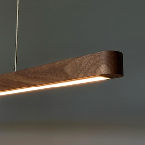 Może przedstawiać: Liniowa lampa wisząca wykonana z ciemnobrązowego drewna z widocznym naturalnym usłojeniem. Wydłużony, prostokątny korpus posiada ciągły zintegrowany pasek świetlny LED wzdłuż dolnej krawędzi, zawieszony na cienkiej metalowej lince.