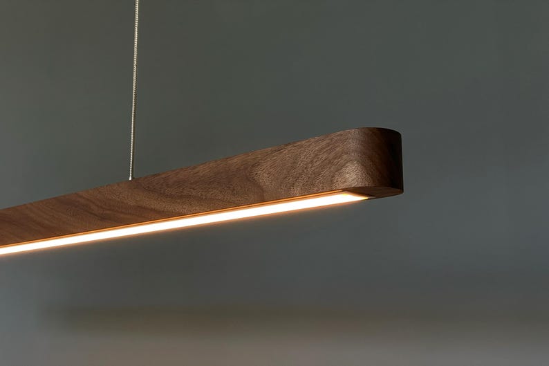 Może przedstawiać: Liniowa lampa wisząca wykonana z ciemnobrązowego drewna z widocznym naturalnym usłojeniem. Wydłużony, prostokątny korpus posiada ciągły zintegrowany pasek świetlny LED wzdłuż dolnej krawędzi, zawieszony na cienkiej metalowej lince.
