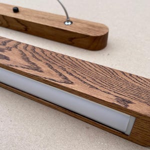 Puede incluir: Una l&aacute;mpara colgante lineal con un marco rectangular de madera con un patr&oacute;n de veta de madera oscura prominente. La l&aacute;mpara incluye un panel difusor de luz blanco esmerilado central y est&aacute; suspendida por cables met&aacute;licos finos con herrajes.