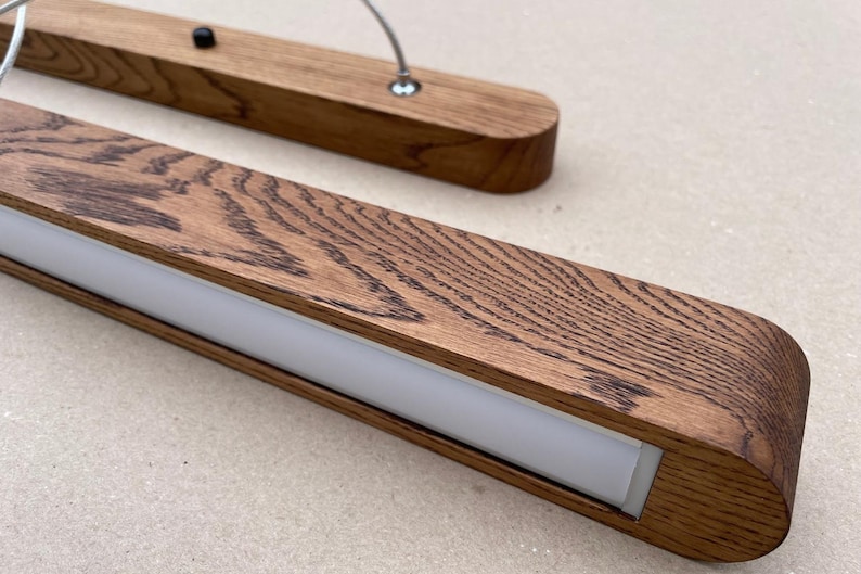 Puede incluir: Una l&aacute;mpara colgante lineal con un marco rectangular de madera con un patr&oacute;n de veta de madera oscura prominente. La l&aacute;mpara incluye un panel difusor de luz blanco esmerilado central y est&aacute; suspendida por cables met&aacute;licos finos con herrajes.