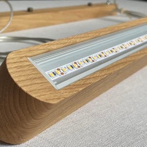 Puede incluir: Una l&aacute;mpara colgante lineal con una carcasa de madera de tono claro natural y un canal de aluminio empotrado que contiene una tira LED visible. La l&aacute;mpara est&aacute; suspendida por cables met&aacute;licos finos unidos a un dosel de techo de madera a juego.