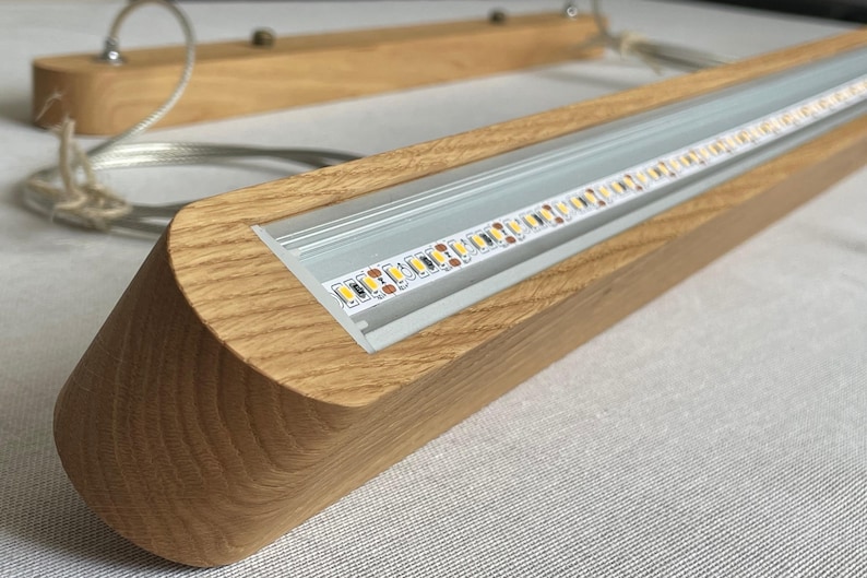 Puede incluir: Una l&aacute;mpara colgante lineal con una carcasa de madera de tono claro natural y un canal de aluminio empotrado que contiene una tira LED visible. La l&aacute;mpara est&aacute; suspendida por cables met&aacute;licos finos unidos a un dosel de techo de madera a juego.