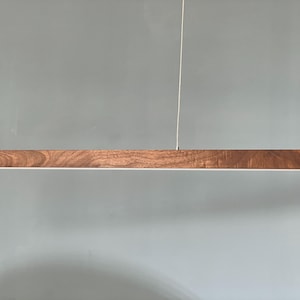Może przedstawiać: Liniowa lampa wisząca składająca się z długiej, poziomej drewnianej belki z widocznym naturalnym usłojeniem. Oprawa jest zawieszona na dw&oacute;ch cienkich, pionowych linkach i posiada biały pasek świetlny zintegrowany z dolną krawędzią.