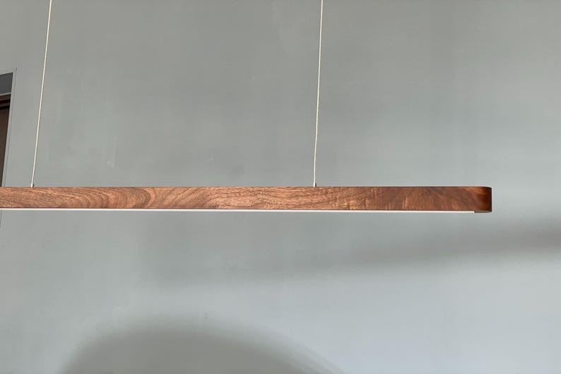 Może przedstawiać: Liniowa lampa wisząca składająca się z długiej, poziomej drewnianej belki z widocznym naturalnym usłojeniem. Oprawa jest zawieszona na dw&oacute;ch cienkich, pionowych linkach i posiada biały pasek świetlny zintegrowany z dolną krawędzią.