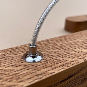 Puede incluir: Una pieza rectangular de madera de roble marr&oacute;n claro con un grano prominente, que presenta un soporte circular de metal cromado unido a la superficie superior. Un cable fino trenzado de color plateado se extiende hacia arriba desde el centro.