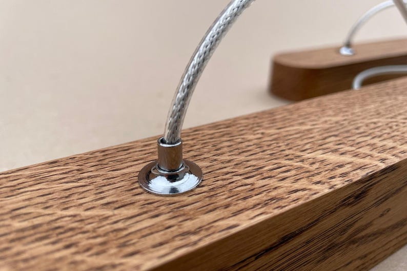 Puede incluir: Una pieza rectangular de madera de roble marr&oacute;n claro con un grano prominente, que presenta un soporte circular de metal cromado unido a la superficie superior. Un cable fino trenzado de color plateado se extiende hacia arriba desde el centro.