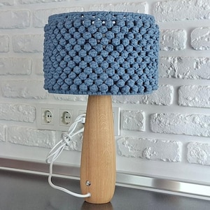 Puede incluir: Una lámpara de mesa artesanal con una pantalla de macramé azul. La lámpara tiene una base de madera y un cable eléctrico blanco. La lámpara está sobre una superficie oscura frente a una pared de ladrillos blancos.