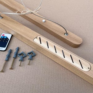Puede incluir: Una l&aacute;mpara lineal de madera clara con un panel central de listones diagonales, un cable de suspensi&oacute;n plateado, un peque&ntilde;o mando a distancia con botones y cuatro tornillos met&aacute;licos con tacos de pl&aacute;stico para su instalaci&oacute;n.