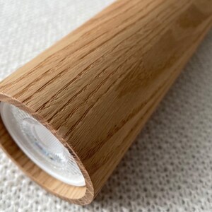 Op de afbeelding: Cilindrische houten lamp met een lichte eikennerf. De lamp heeft een afgerond uiteinde met een heldere witte lichtbron. De houtnerf is zichtbaar op het oppervlak.