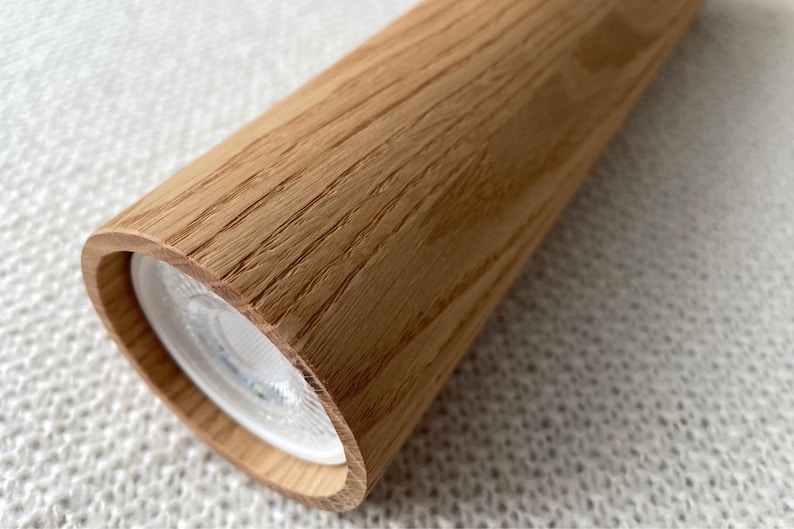 Op de afbeelding: Cilindrische houten lamp met een lichte eikennerf. De lamp heeft een afgerond uiteinde met een heldere witte lichtbron. De houtnerf is zichtbaar op het oppervlak.