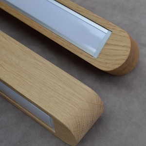 Puede incluir: Dos luminarias lineales fabricadas en madera de tono claro con un patr&oacute;n de veta visible. Cada luminaria cuenta con un panel difusor blanco rectangular largo insertado en el centro del marco de madera, con extremos redondeados.