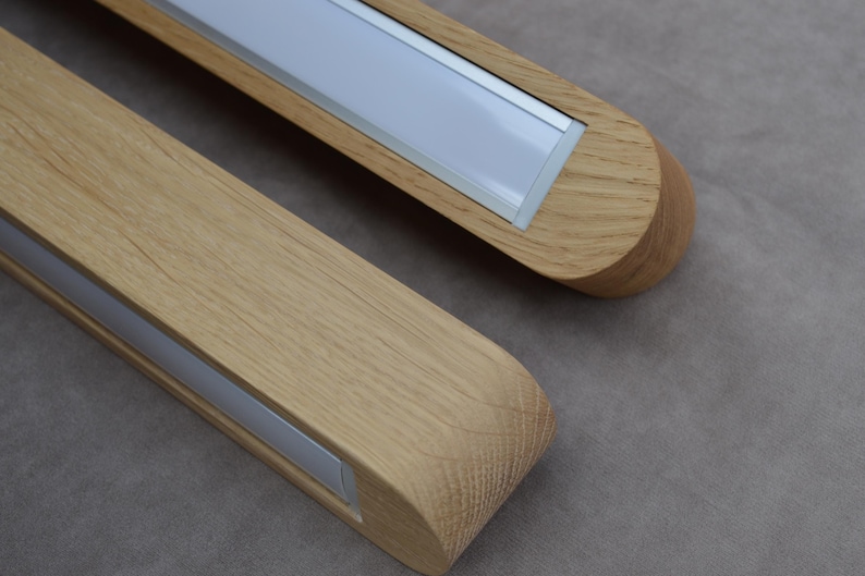 Puede incluir: Dos luminarias lineales fabricadas en madera de tono claro con un patr&oacute;n de veta visible. Cada luminaria cuenta con un panel difusor blanco rectangular largo insertado en el centro del marco de madera, con extremos redondeados.