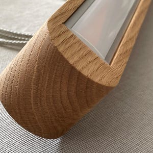 Puede incluir: Una luminaria lineal con una carcasa de madera de color claro natural con un patr&oacute;n de veta visible. La luminaria incluye un panel difusor blanco empotrado y est&aacute; conectada a un cable met&aacute;lico plateado fino que se extiende por el lateral.