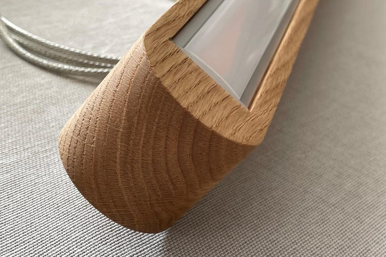 Puede incluir: Una luminaria lineal con una carcasa de madera de color claro natural con un patr&oacute;n de veta visible. La luminaria incluye un panel difusor blanco empotrado y est&aacute; conectada a un cable met&aacute;lico plateado fino que se extiende por el lateral.