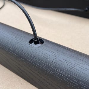 Puede incluir: Primer plano de un componente de madera negro para una l&aacute;mpara con un agujero para un cable. La veta de la madera es visible. Un cable negro est&aacute; insertado en el agujero. Otro componente similar est&aacute; en el fondo.