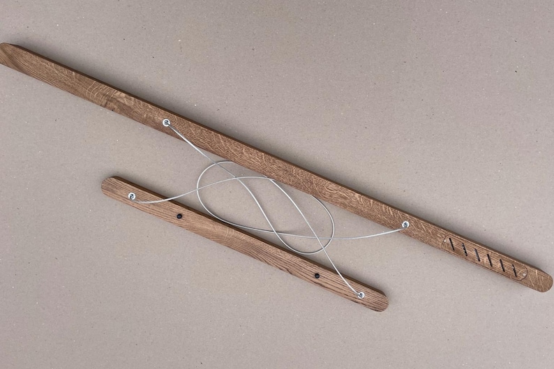 Puede incluir: Dos listones de madera alargados de color marr&oacute;n medio con una textura de veta visible. Los listones est&aacute;n conectados por un alambre met&aacute;lico fino de color plateado que forma un bucle entre ellos. Un list&oacute;n tiene ranuras diagonales.
