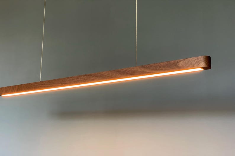 Może przedstawiać: Liniowa lampa wisząca wykonana z ciemnobrązowego drewna o widocznym usłojeniu. Oprawa ma długi, wąski, prostokątny kształt zawieszony na dw&oacute;ch cienkich przewodach, emitujący ciepłe, jasne światło z dolnej części urządzenia.