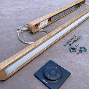 Puede incluir: Luminaria LED de madera con una tira de luz blanca, un mando a distancia y accesorios de montaje. La luminaria es de madera de color claro y tiene un dise&ntilde;o moderno. El mando a distancia es negro con un panel de control circular.