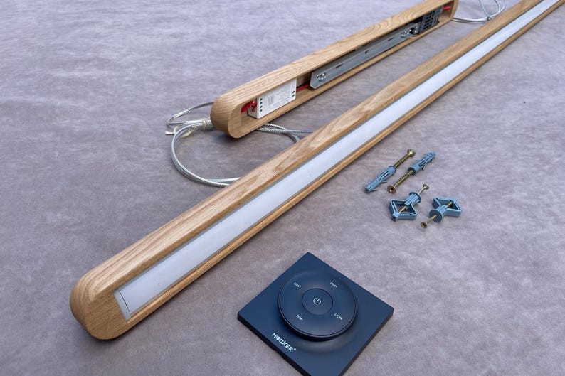 Puede incluir: Luminaria LED de madera con una tira de luz blanca, un mando a distancia y accesorios de montaje. La luminaria es de madera de color claro y tiene un dise&ntilde;o moderno. El mando a distancia es negro con un panel de control circular.