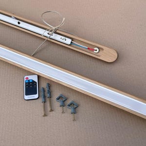 Puede incluir: Una l&aacute;mpara lineal con marco de madera clara y difusor blanco. El conjunto incluye un mando a distancia con botones y cuatro tornillos de montaje met&aacute;licos con tacos de pl&aacute;stico gris para su instalaci&oacute;n en pared o techo.