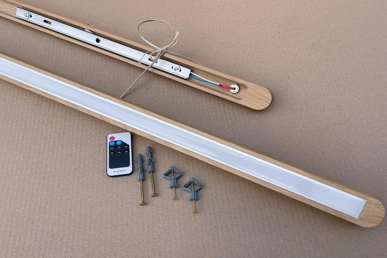 Puede incluir: Una l&aacute;mpara lineal con marco de madera clara y difusor blanco. El conjunto incluye un mando a distancia con botones y cuatro tornillos de montaje met&aacute;licos con tacos de pl&aacute;stico gris para su instalaci&oacute;n en pared o techo.