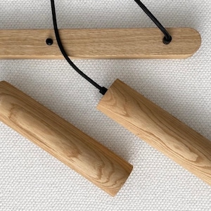 Op de afbeelding: Houten hanglamp met twee cilindrische kappen. De kappen zijn gemaakt van lichtgekleurd hout en hangen aan zwarte snoeren die aan een houten balk zijn bevestigd. De armatuur is gemonteerd op een wit, getextureerd oppervlak.