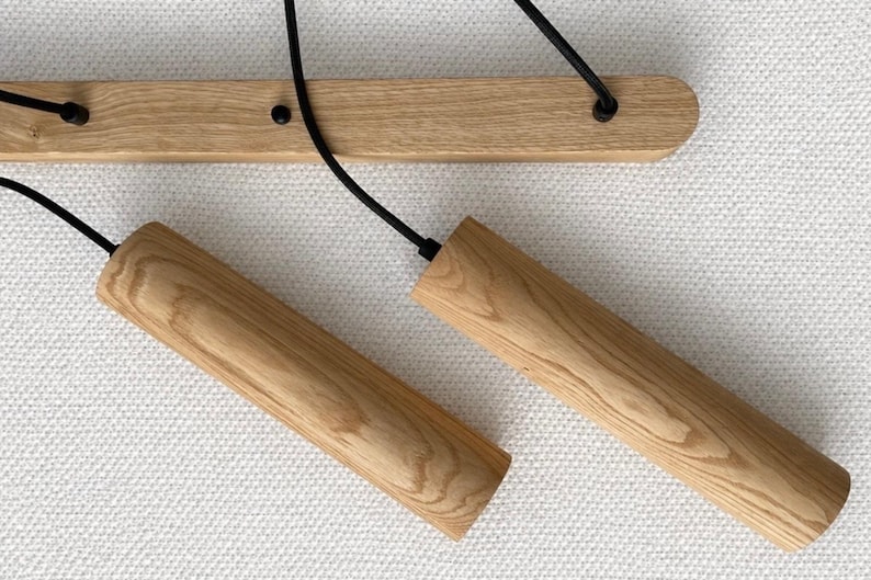 Op de afbeelding: Houten hanglamp met twee cilindrische kappen. De kappen zijn gemaakt van lichtgekleurd hout en hangen aan zwarte snoeren die aan een houten balk zijn bevestigd. De armatuur is gemonteerd op een wit, getextureerd oppervlak.