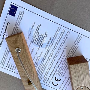 Puede incluir: Dos bloques rectangulares de madera clara con veta visible, cada uno con un peque&ntilde;o componente circular met&aacute;lico y un cord&oacute;n fino, est&aacute;n colocados sobre un documento blanco titulado Declaration of Conformity de la Uni&oacute;n Europea.