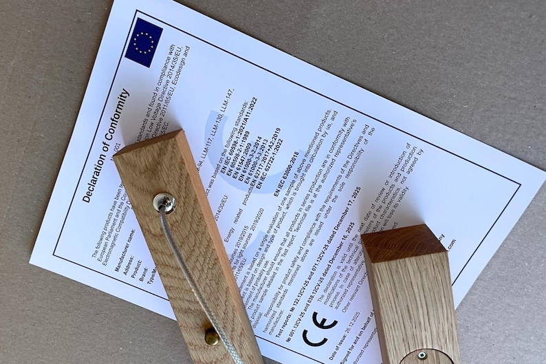 Puede incluir: Dos bloques rectangulares de madera clara con veta visible, cada uno con un peque&ntilde;o componente circular met&aacute;lico y un cord&oacute;n fino, est&aacute;n colocados sobre un documento blanco titulado Declaration of Conformity de la Uni&oacute;n Europea.