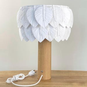 May include: Table lamp featuring a white feather-shaped lampshade and a wooden base. The lamp has a cylindrical wooden base and a white cord with a plug. The shade is made of layered white feather-like elements. The lamp is on a wooden table.