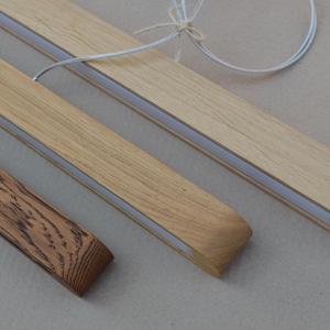 Puede incluir: Tres luminarias lineales de madera con acabado liso. Dos luminarias son de madera clara con veta visible y una tira de luz central blanca. Una luminaria es de madera oscura y texturizada con un patr&oacute;n de veta muy marcado.