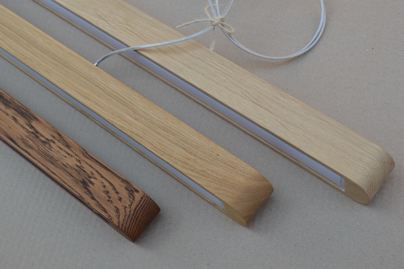 Puede incluir: Tres luminarias lineales de madera con acabado liso. Dos luminarias son de madera clara con veta visible y una tira de luz central blanca. Una luminaria es de madera oscura y texturizada con un patr&oacute;n de veta muy marcado.