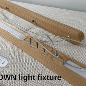 Puede incluir: Una l&aacute;mpara lineal de madera con acabado de veta clara, que cuenta con paneles LED integrados, cables de suspensi&oacute;n plateados y un peque&ntilde;o control remoto blanco. La l&aacute;mpara incluye el texto UP/DOWN light fixture.