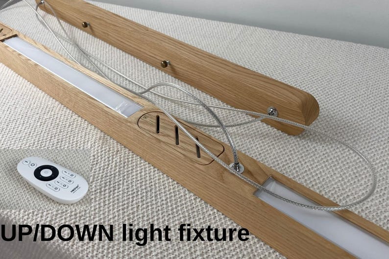 Puede incluir: Una l&aacute;mpara lineal de madera con acabado de veta clara, que cuenta con paneles LED integrados, cables de suspensi&oacute;n plateados y un peque&ntilde;o control remoto blanco. La l&aacute;mpara incluye el texto UP/DOWN light fixture.