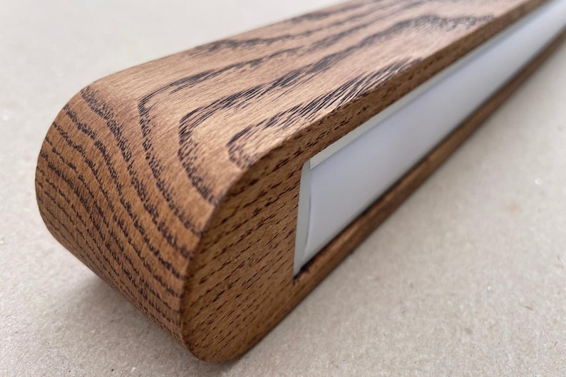 Puede incluir: Una luminaria lineal con una carcasa de madera marr&oacute;n natural con un patr&oacute;n de veta prominente y un extremo redondeado. La luminaria incluye una tira difusora blanca larga y empotrada a lo largo de su longitud para emitir luz de forma uniforme.