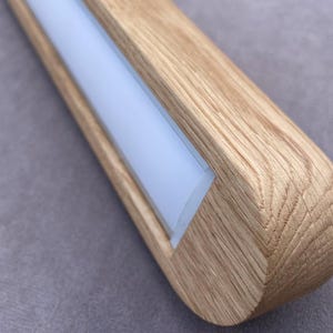 Puede incluir: Primer plano de una l&aacute;mpara de madera con un extremo redondeado. La l&aacute;mpara tiene una veta de madera clara y un panel de luz central rectangular blanco esmerilado. La l&aacute;mpara est&aacute; sobre un fondo gris.