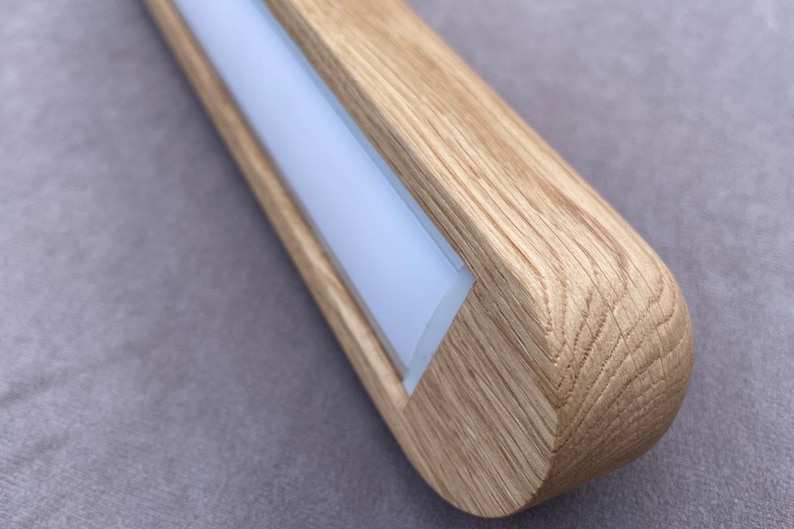 Puede incluir: Primer plano de una l&aacute;mpara de madera con un extremo redondeado. La l&aacute;mpara tiene una veta de madera clara y un panel de luz central rectangular blanco esmerilado. La l&aacute;mpara est&aacute; sobre un fondo gris.