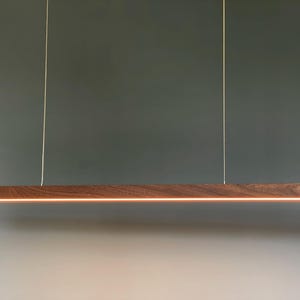 Może przedstawiać: Liniowa lampa wisząca składająca się z długiej, prostokątnej belki z ciemnobrązowego drewna o widocznym usłojeniu. Oprawa jest zawieszona na dw&oacute;ch cienkich, pionowych linkach i emituje ciepłe, ciągłe pasmo światła od spodu.