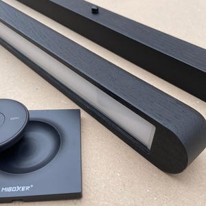 Puede incluir: Luminarias LED de madera negra con mando a distancia y base. Las luminarias tienen forma rectangular con una tira de luz empotrada. El mando a distancia y la base son negros y tienen la marca "MIBOXER".