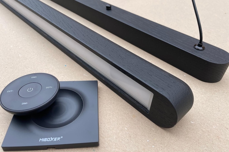 Puede incluir: Luminarias LED de madera negra con mando a distancia y base. Las luminarias tienen forma rectangular con una tira de luz empotrada. El mando a distancia y la base son negros y tienen la marca "MIBOXER".