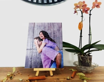 Puede incluir: Una pareja posa para una foto, la mujer lleva al hombre a cuestas. La foto está impresa en un rompecabezas, que se muestra en un pequeño caballete de madera. El rompecabezas está sobre una mesa de madera con flores secas y palitos de canela.