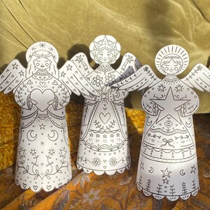 Op de afbeelding: Drie witte papieren engelenfiguren met gedetailleerde zwarte lijntekeningen. Elke engel heeft vleugels, een halo en een patroonjurk. De engelen zijn versierd met harten, sterren, manen en bloemmotieven, perfect om in te kleuren of te knutselen.