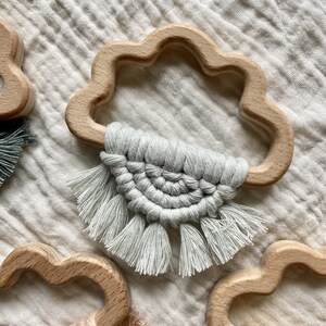 K&ouml;nnte beinhalten: Ein Set aus drei Holz-Bei&szlig;spielzeugen in Wolkenform, jedes mit einem anderen farbigen Makramee-Quaste. Die Quasten sind in verschiedenen Farben gefertigt, darunter Grau, Beige und Gelb.