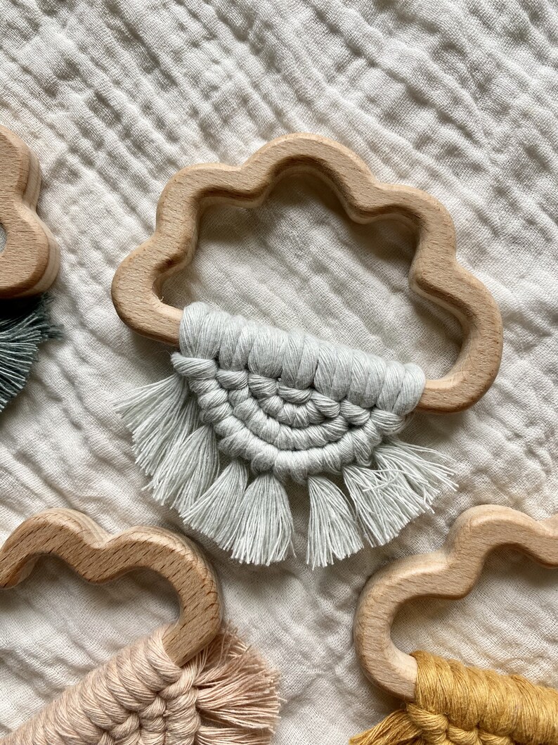 K&ouml;nnte beinhalten: Ein Set aus drei Holz-Bei&szlig;spielzeugen in Wolkenform, jedes mit einem anderen farbigen Makramee-Quaste. Die Quasten sind in verschiedenen Farben gefertigt, darunter Grau, Beige und Gelb.
