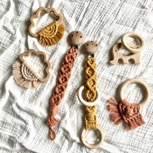 K&ouml;nnte beinhalten: Ein Set aus Holz-Bei&szlig;ringen mit bunten Makramee- und Fransen-Akzenten. Die Bei&szlig;ringe sind in Form einer Wolke, eines Elefanten und zweier Ringe. Die Bei&szlig;ringe sind perfekt f&uuml;r Babys zum Kauen und Erkunden.