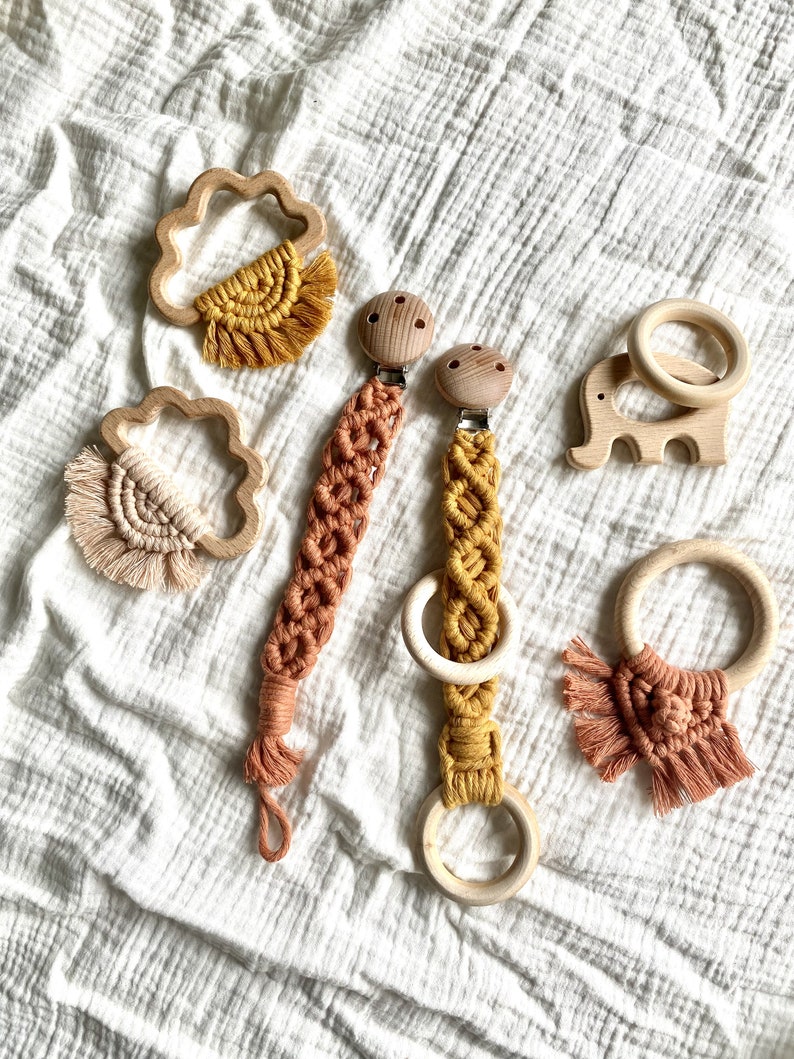 K&ouml;nnte beinhalten: Ein Set aus Holz-Bei&szlig;ringen mit bunten Makramee- und Fransen-Akzenten. Die Bei&szlig;ringe sind in Form einer Wolke, eines Elefanten und zweier Ringe. Die Bei&szlig;ringe sind perfekt f&uuml;r Babys zum Kauen und Erkunden.