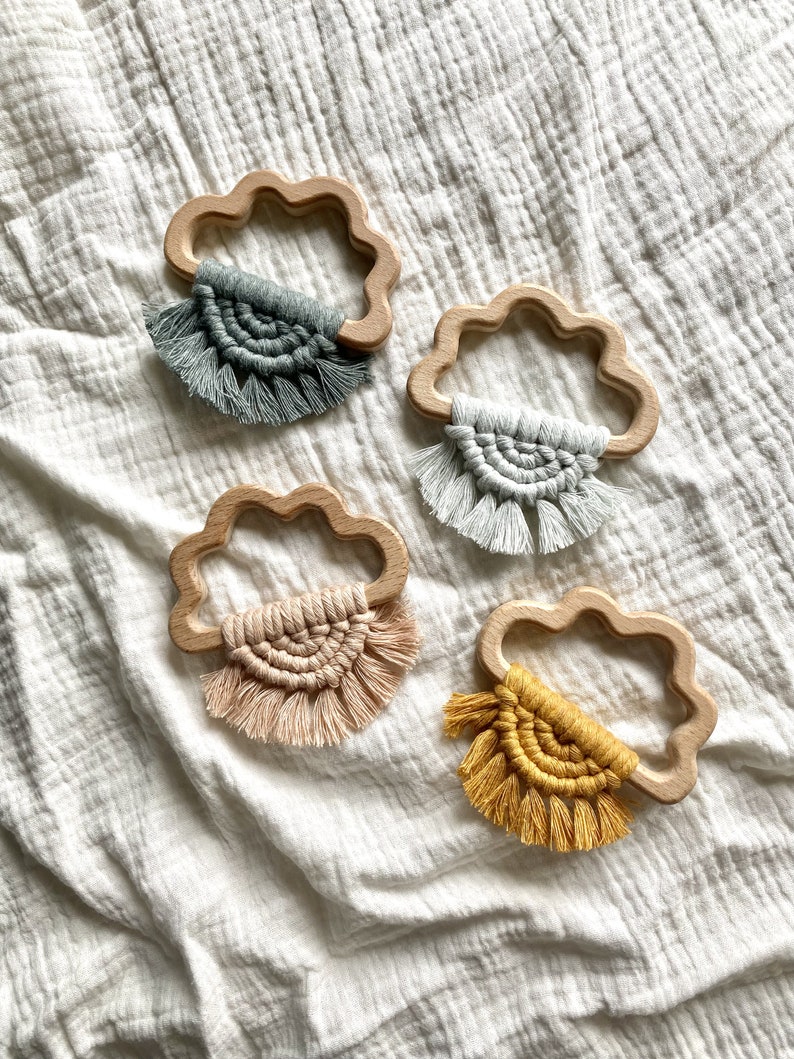 K&ouml;nnte beinhalten: Vier h&ouml;lzerne Bei&szlig;ringe in Wolkenform mit verschiedenen bunten geh&auml;kelten Quasten. Die Quasten sind in den Farben Grau, Beige, Rosa und Gelb.