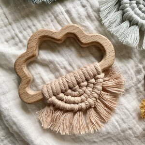 K&ouml;nnte beinhalten: Ein wolkenf&ouml;rmiger Bei&szlig;ring aus Holz mit einem rosafarbenen Makramee-Quaste, der daran h&auml;ngt. Der Bei&szlig;ring liegt auf einem wei&szlig;en Stoffhintergrund.