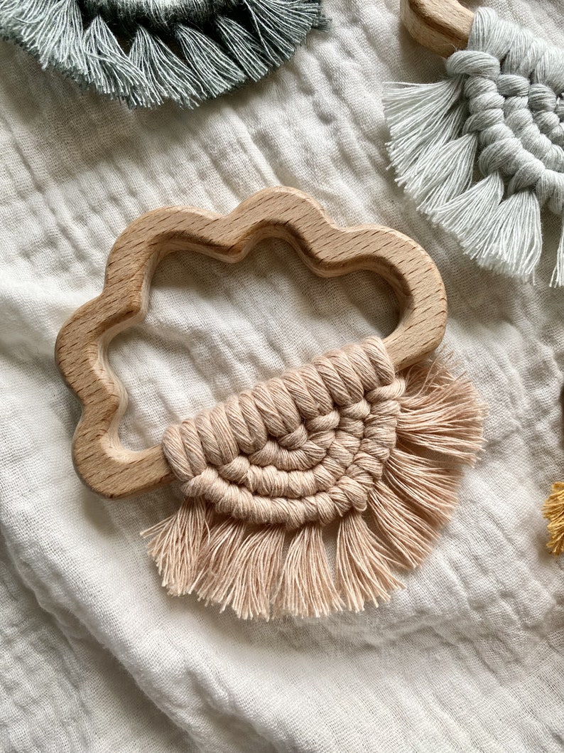 K&ouml;nnte beinhalten: Ein wolkenf&ouml;rmiger Bei&szlig;ring aus Holz mit einem rosafarbenen Makramee-Quaste, der daran h&auml;ngt. Der Bei&szlig;ring liegt auf einem wei&szlig;en Stoffhintergrund.