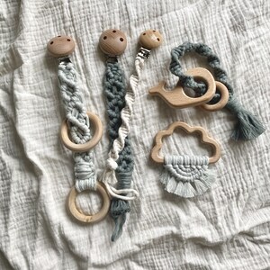 K&ouml;nnte beinhalten: Vier Holz-Bei&szlig;ringe mit Makramee-B&auml;ndern und Clips. Die Bei&szlig;ringe sind in Form eines Rings, einer Wolke, eines Wals und einer Spirale. Die Bei&szlig;ringe sind in Graut&ouml;nen und Wei&szlig;.